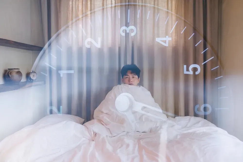 A man sits up in bed wrapped in a duvet, with a large clock face double-exposed over the scene, its hand sweeping across the numbers.