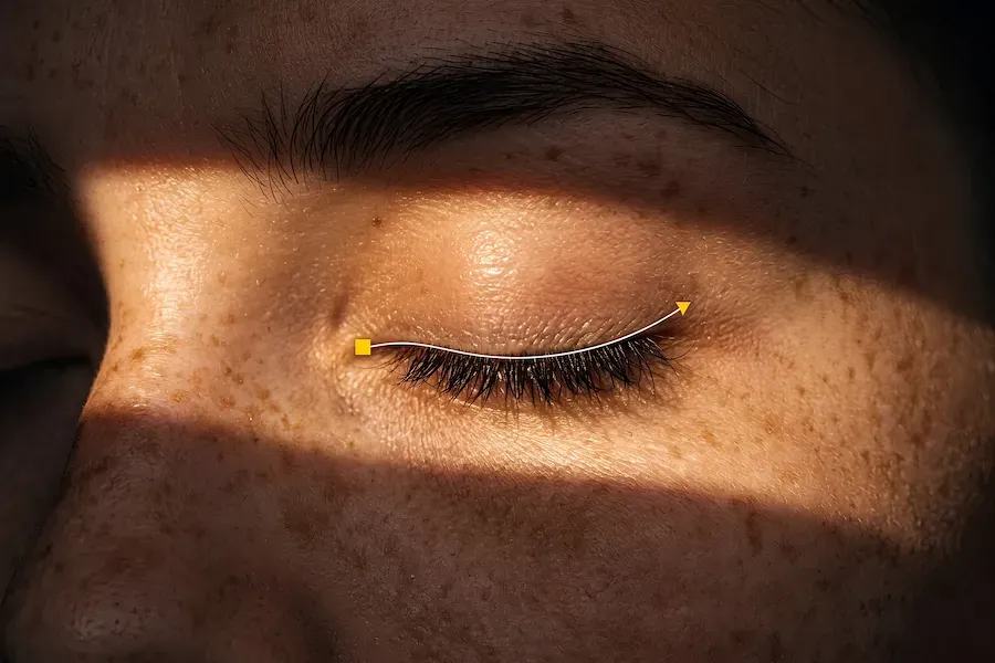 Extreme close-up of a closed eye and freckled skin with a hard shaft of light falling across the face, and a thin yellow line traced along the lower lashes.