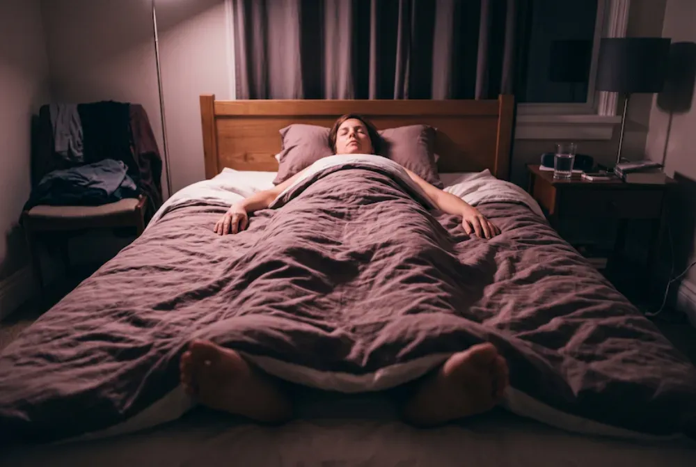 Woman lying on her back in bed at night, seen from the foot of the bed looking up the length of her body toward the headboard.