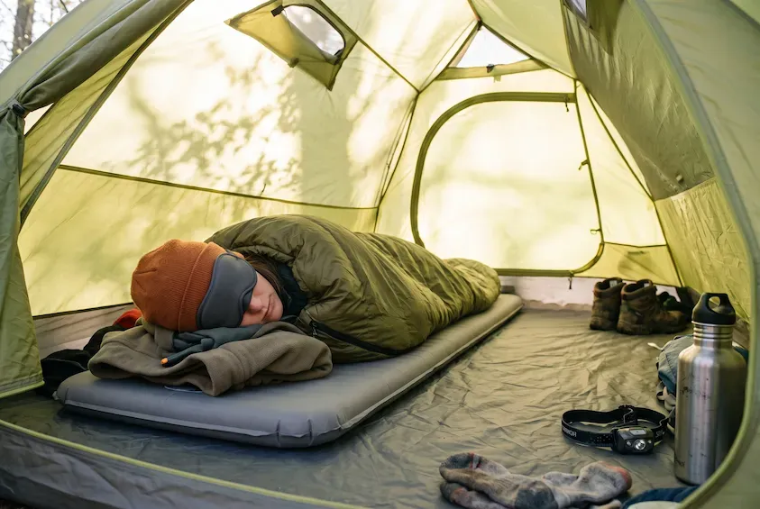 Person asleep in a sleeping bag inside a sunlit tent, wearing a sleep mask and woolly hat, on an inflatable mat over a foam mat.