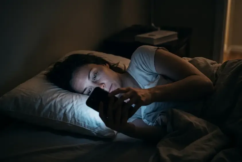 A woman lying in a dark bedroom, her face illuminated by the cool glow of the smartphone she is holding.