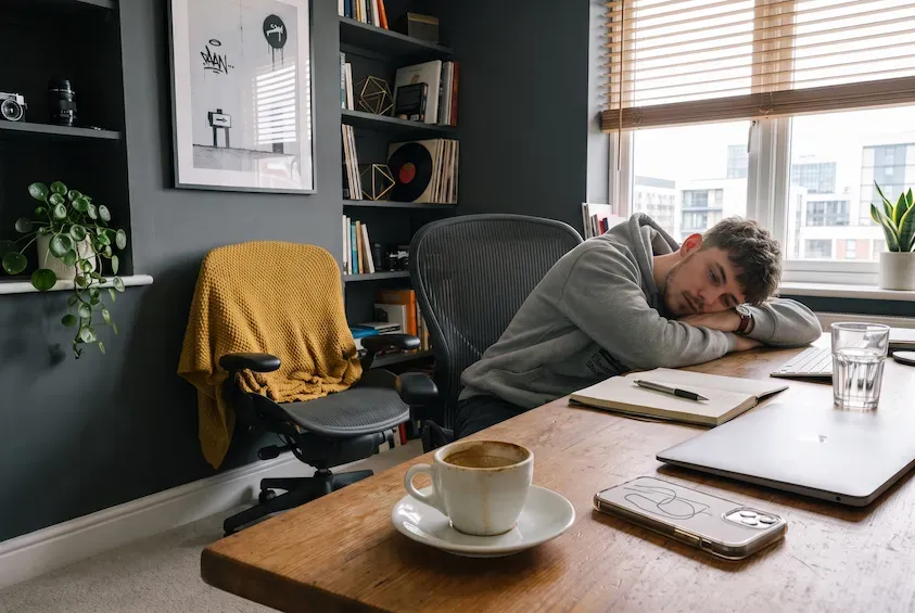 Man asleep with his head on his arms at a home-office desk, a finished cup of coffee and a closed laptop beside him.