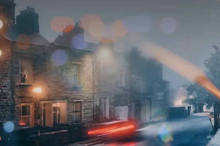A dreamlike, blurred night view of a stone terraced street lit by glowing streetlamps, with a figure visible at one lit window, evoking an unfamiliar place.