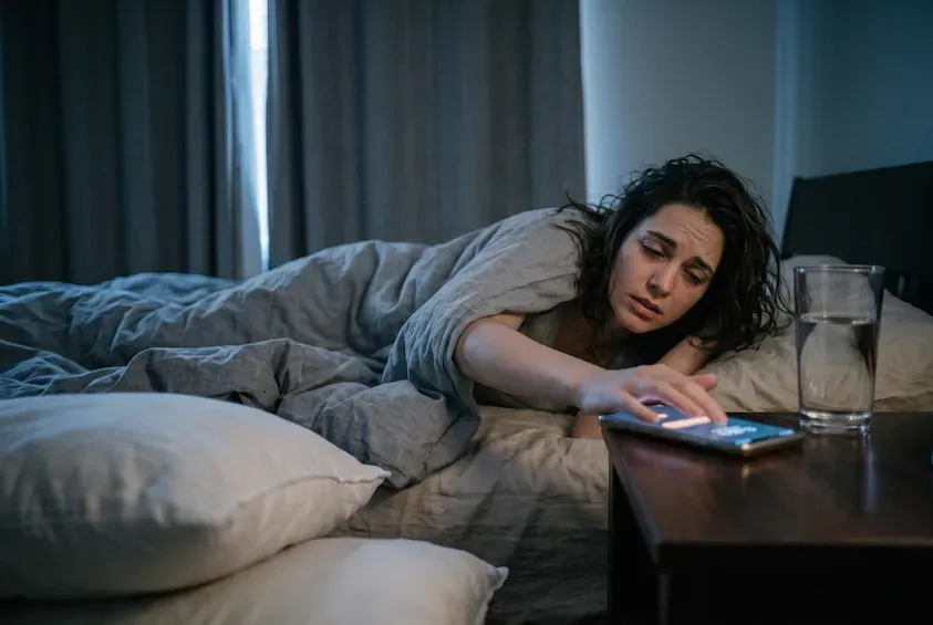 Woman reaching from under the duvet to silence a phone alarm on the bedside table, eyes barely open.