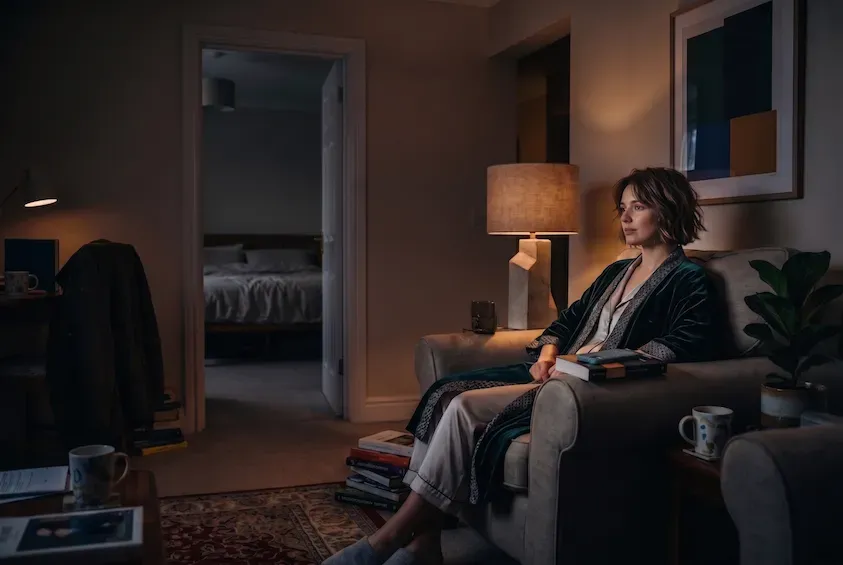 Woman sitting up in an armchair in a lamplit living room in the small hours, a book in her lap and the dark bedroom visible through the doorway.