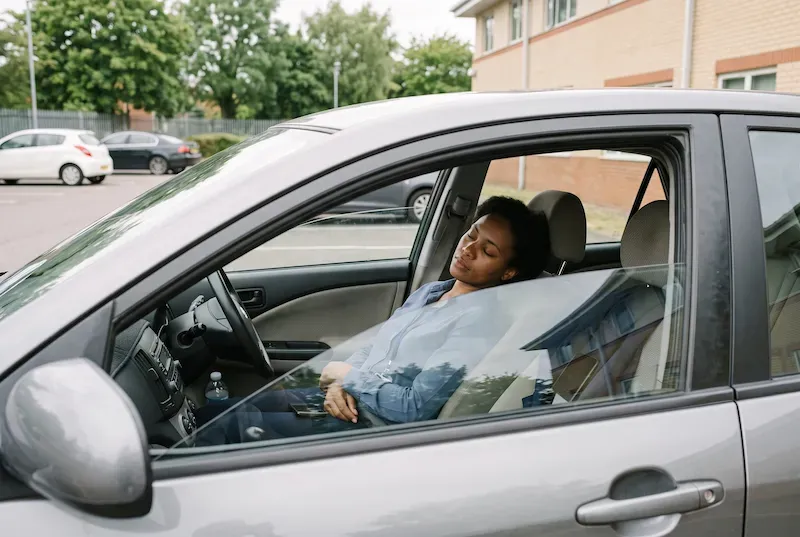 Woman napping in the reclined driver's seat of a parked car in a workplace car park, still in office clothes.