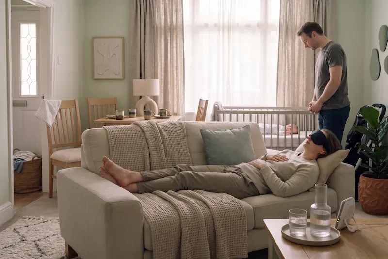 Woman napping on the sofa in a sleep mask while her partner stands by the cot where the baby is sleeping.