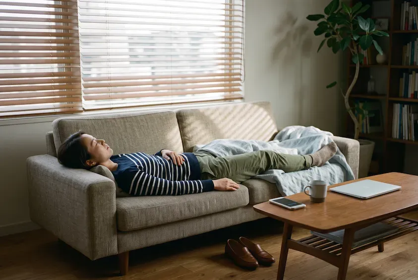 Woman lying flat on a sofa taking a short afternoon nap in daytime clothes, shoes off, blinds half open behind her.