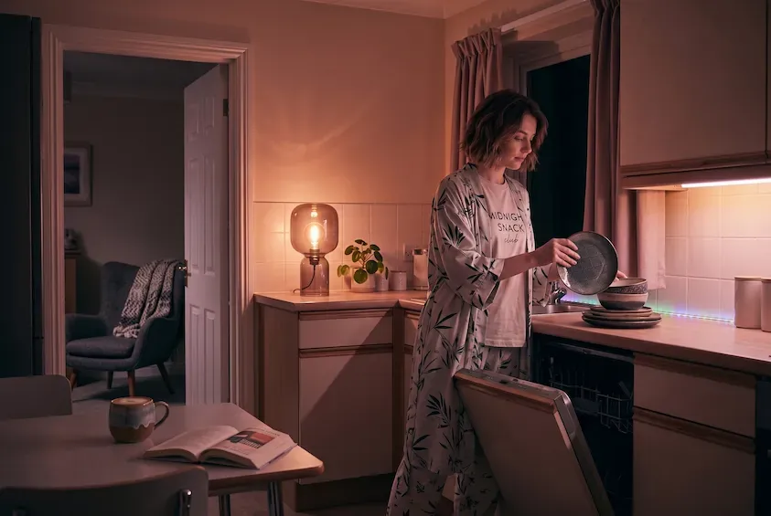 Woman in pyjamas unloading the dishwasher late at night in a lamplit kitchen, an abandoned chair and blanket visible in the room behind.