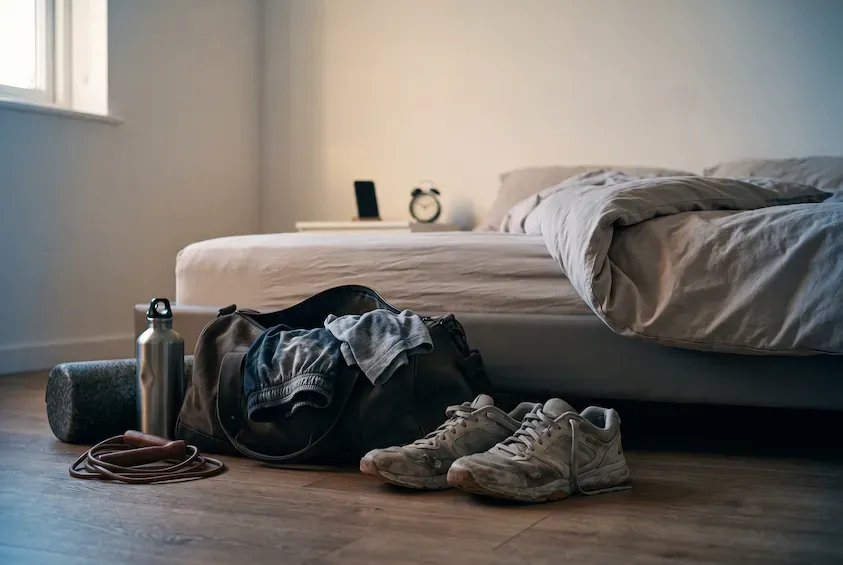 Used gym bag, worn trainers, foam roller and water bottle on the floor beside a turned-back bed.