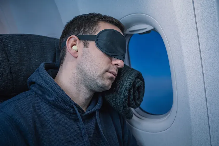 Man asleep in an aeroplane window seat wearing a sleep mask and earplugs, head resting on a rolled jumper against the cabin wall.