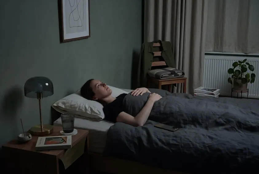 Woman lying awake on her back in a dim green bedroom, eyes open, completely still under the covers.