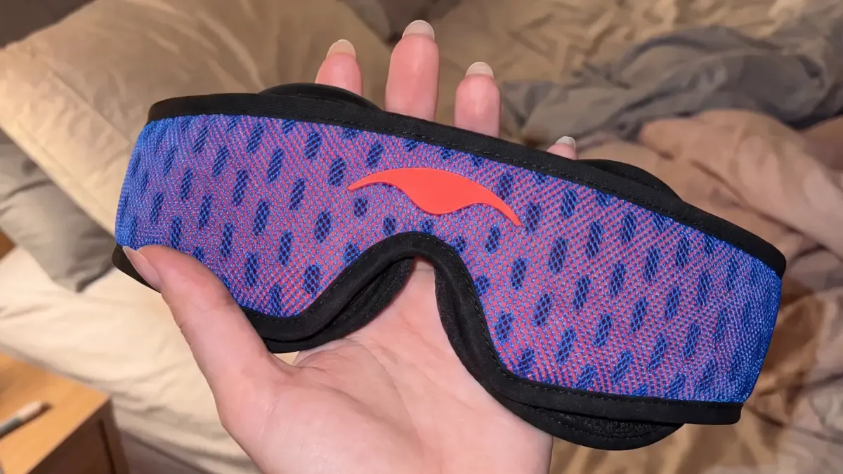 A hand holding Manta PRO Sleep Mask with the bed in the background.