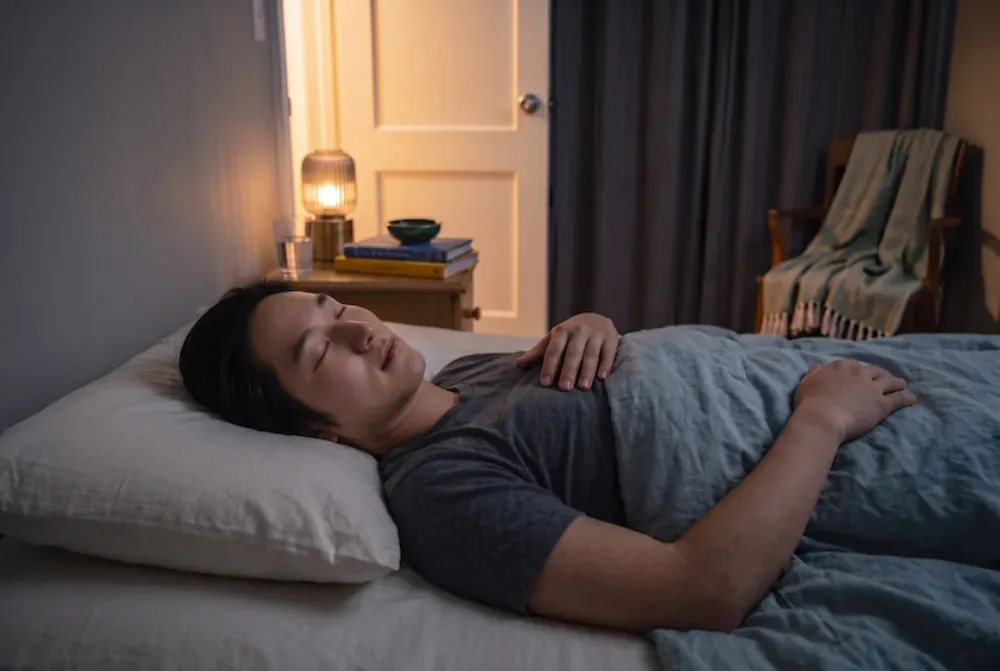 Man lying on his back in bed at night with his eyes closed, one hand resting on his chest, a lamp glowing softly on the bedside table.