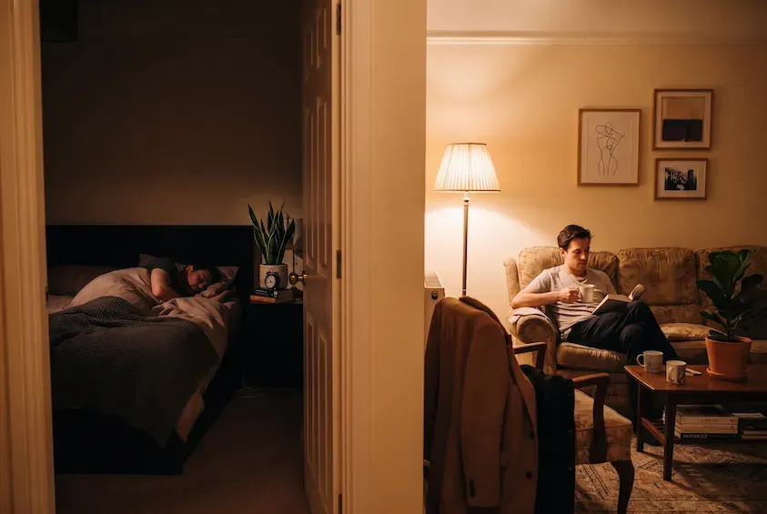 One person asleep in a dark bedroom while another sits reading under a lamp in the lit room next door, seen through the doorway.